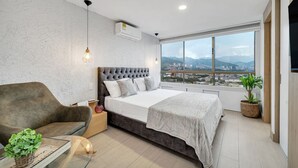 A modern bedroom with a large bed, a sofa, a view of a cityscape, and a large window. - Alcazar Oviedo - 1103 (Medellín)