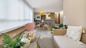 A modern living room with a sofa, dining table, and chairs, a kitchen area in the background, and a large window. - Alcazar Oviedo - 1103 (Medellín)