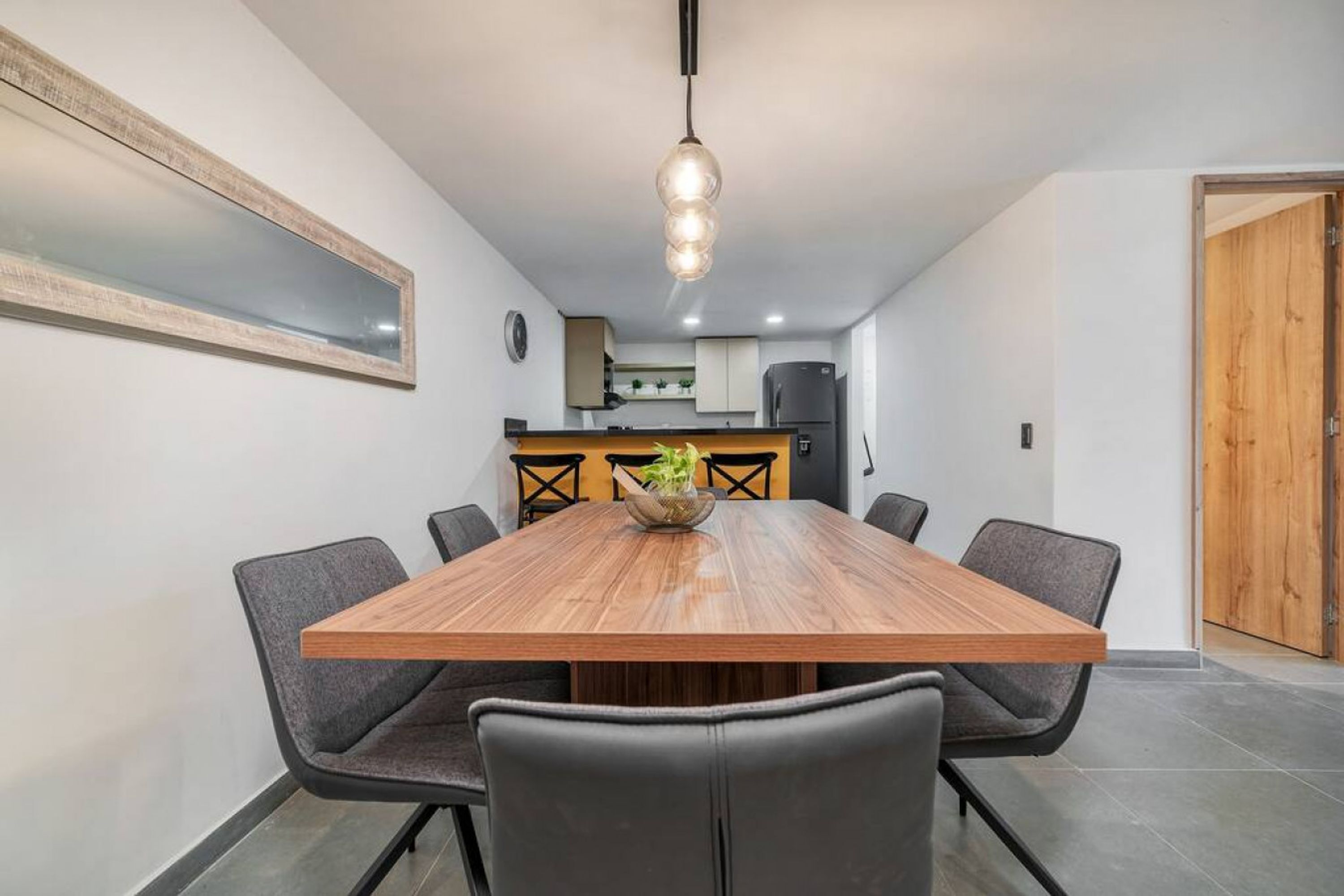 Een eetruimte met een houten tafel, zes stoelen, een hanglamp, een keuken op de achtergrond en een houten deur.