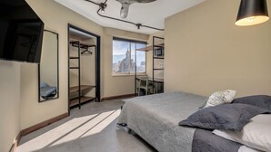 Ett sovrum med en säng, en takfläkt, ett skrivbord, en stol, en garderob och ett stort fönster med utsikt över staden.