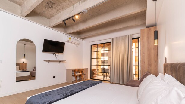Une chambre d’hôtel moderne dotée d’un grand lit, d’un bureau, d’une chaise, d’une télévision et d’une porte-fenêtre coulissante donnant sur un balcon.