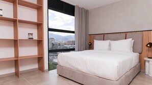 A modern bedroom with a large bed, a wooden bookshelf, and a city view through the window. - Metropolitan Living - 515a (Medellín)