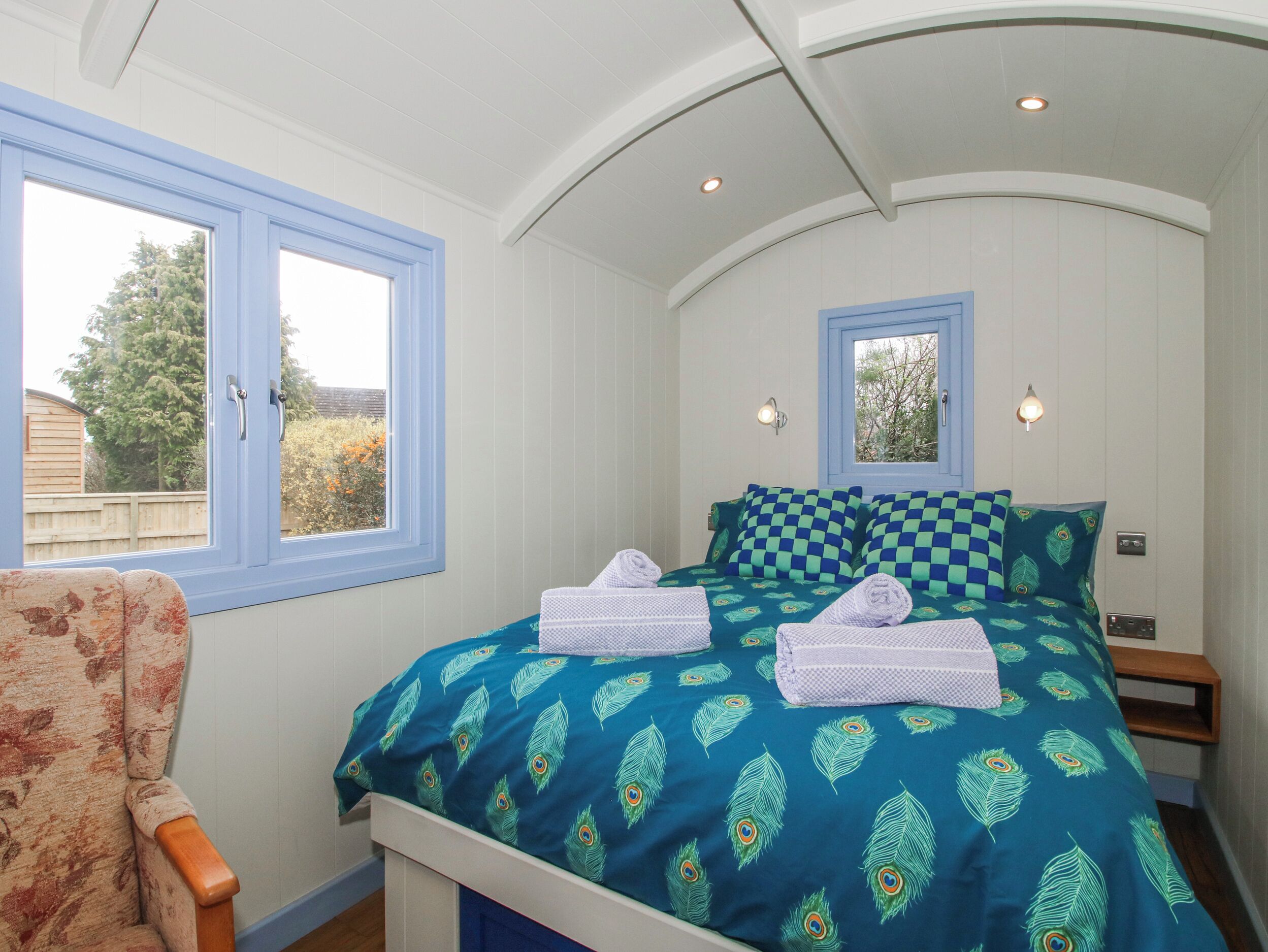 A bedroom with a bed, two windows, a chair, and a small bedside table.