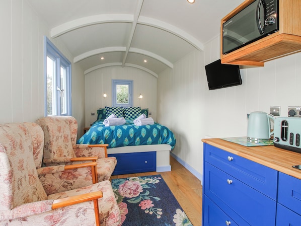 A compact room with a bed, a seating area with floral-patterned chairs, a wooden cabinet, and a mounted television. - Skye (Ludlow)