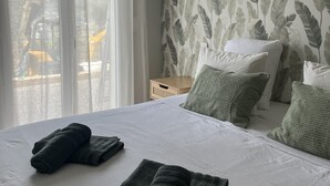 Quarto com cama, almofadas, mesa de cabeceira, abajur e janela com cortinas.