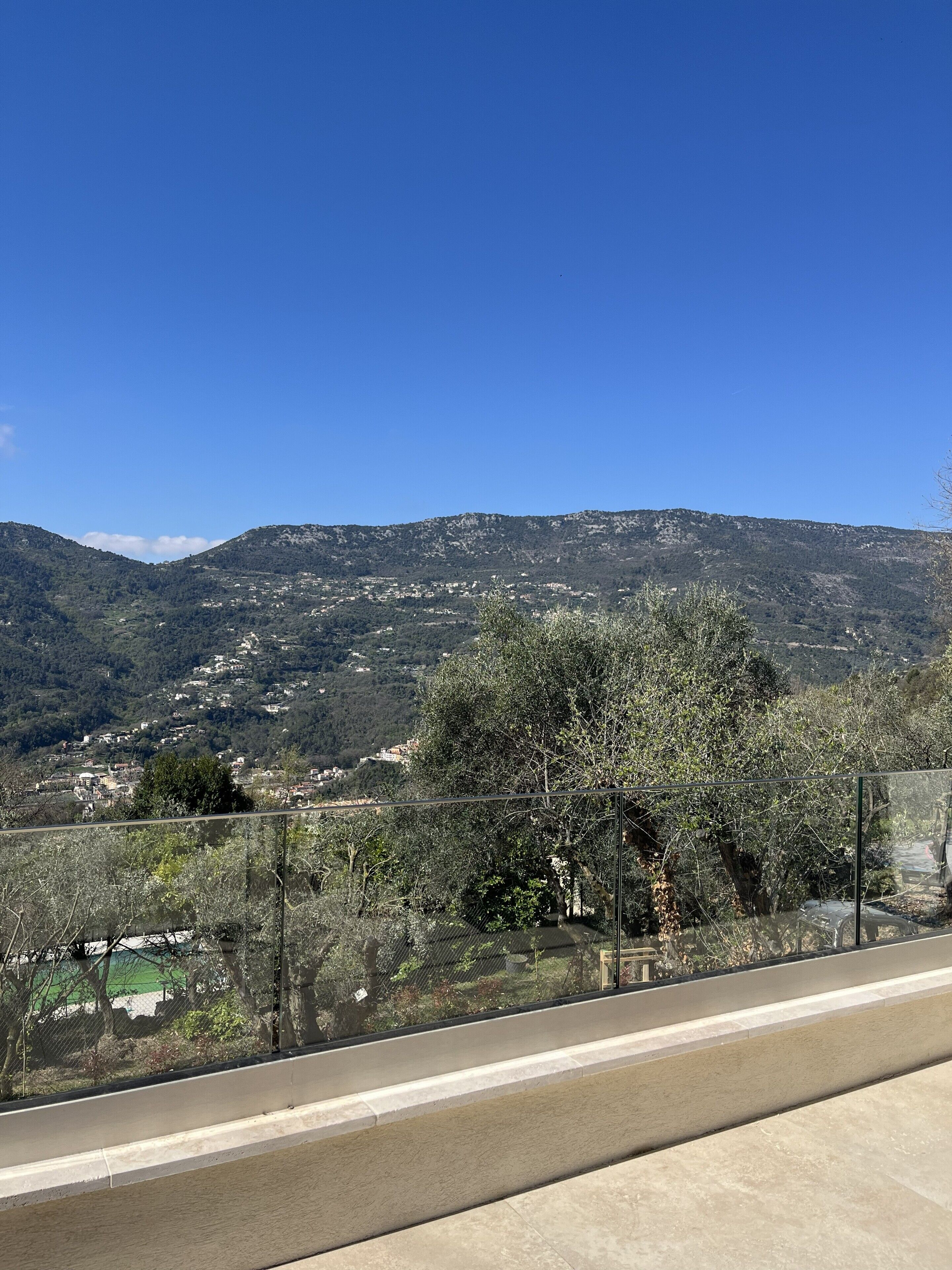 A view from a balcony with a metal railing, overlooking a valley with scattered buildings and hills in the background.