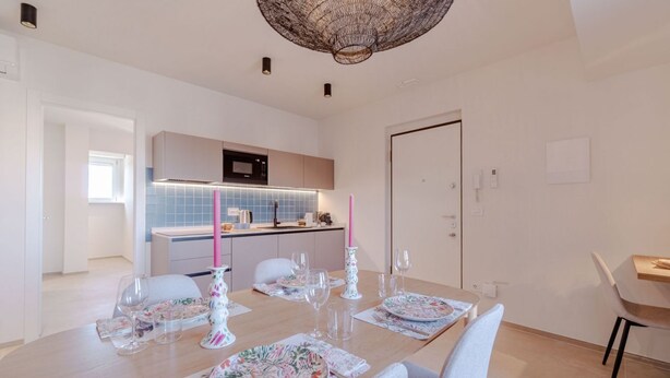 Zona pranzo moderna con tavolo imbandito per quattro persone, cucina sullo sfondo e una porta a destra.