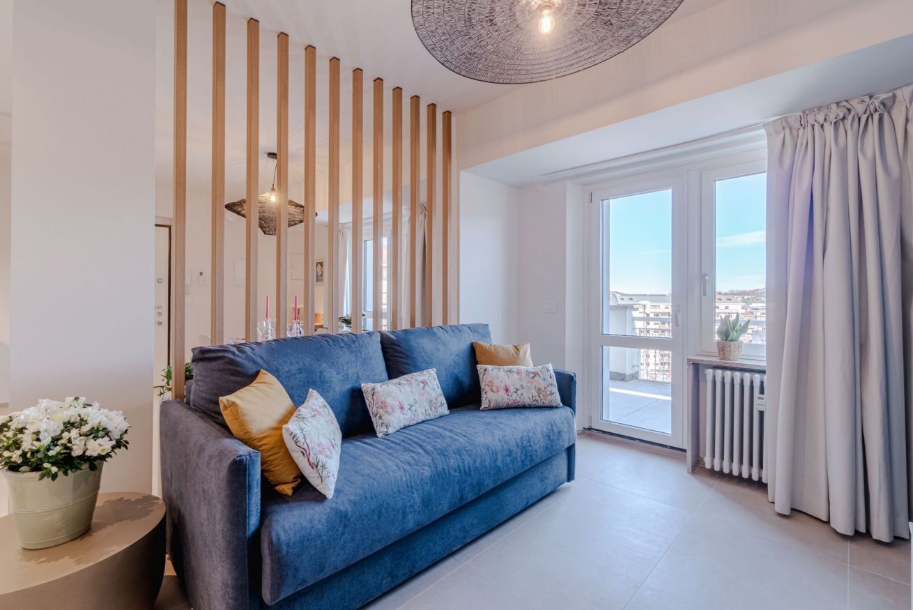 Ein modernes Wohnzimmer mit einem blauen Sofa, dekorativen Kissen, einer Topfpflanze und Blick durch große Fenster.