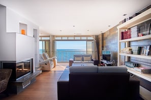 Un salon moderne doté d’une cheminée, de sièges confortables, d’une bibliothèque et d’une grande fenêtre offrant une vue sur la mer.