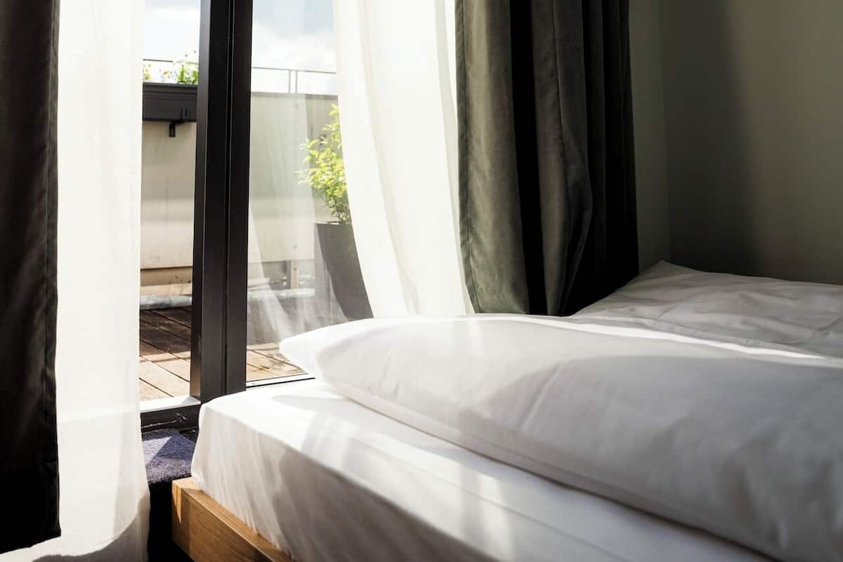 Quarto com cama, cortinas leves e vista para o exterior.