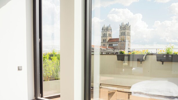 A modern hotel room with a large window, a bed, and a view of a historic building. - ALPAMI Glockenbach  (Munich)