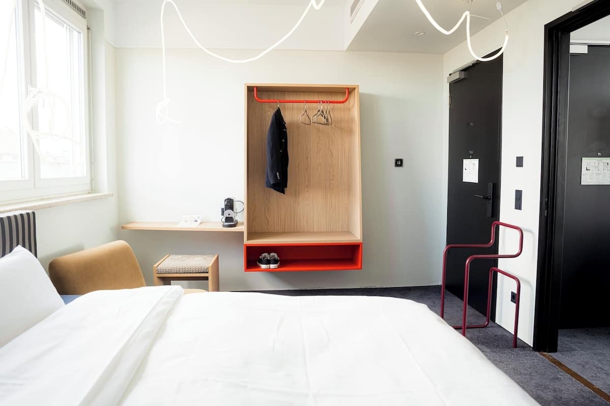 Quarto de hotel com uma cama, uma escrivaninha com cafeteira, um guarda-roupa de madeira, uma cadeira vermelha e uma porta preta.