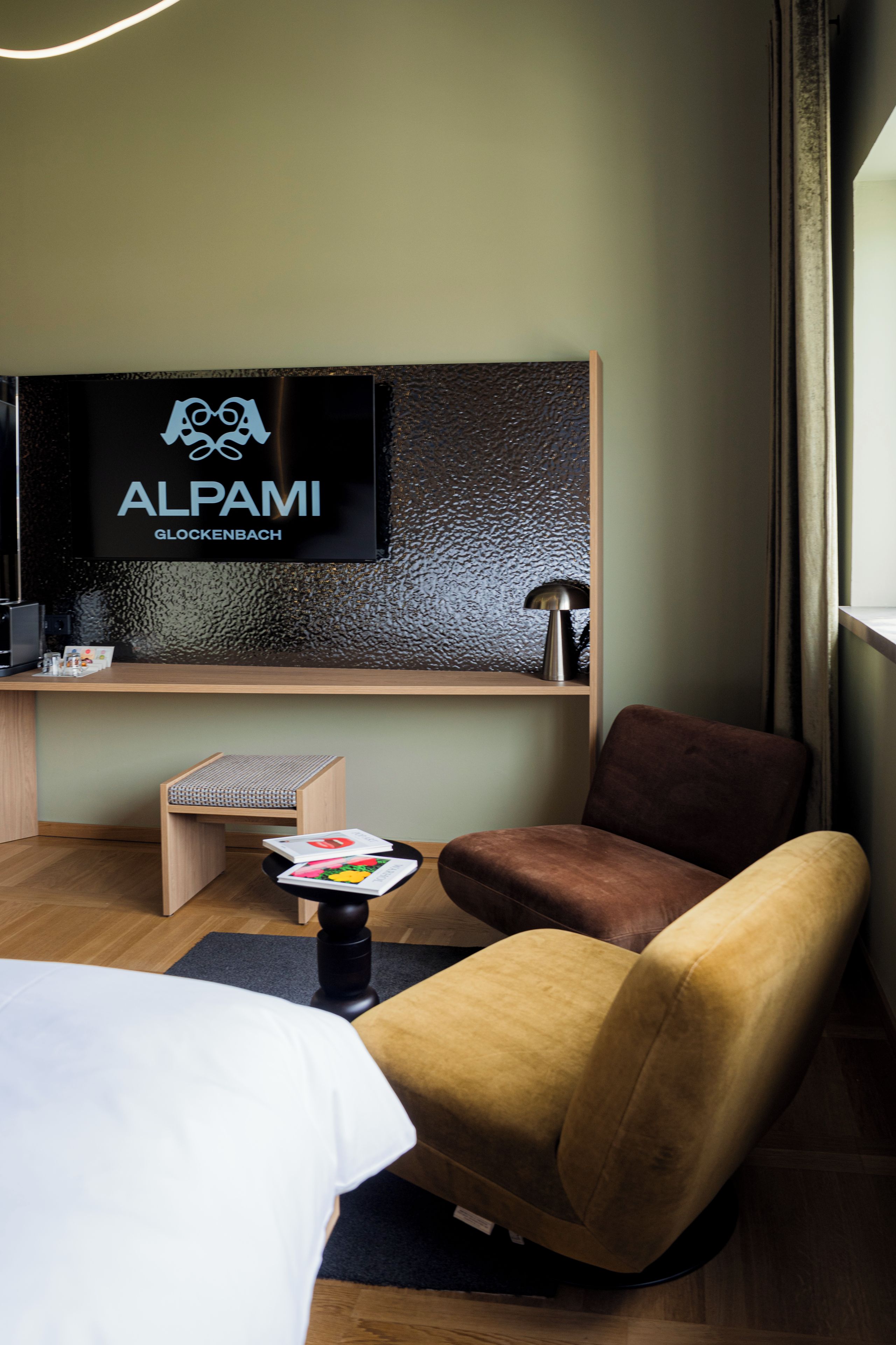 Quarto de hotel com cama, escrivaninha, cadeira, televisão e uma pequena mesa com livros.