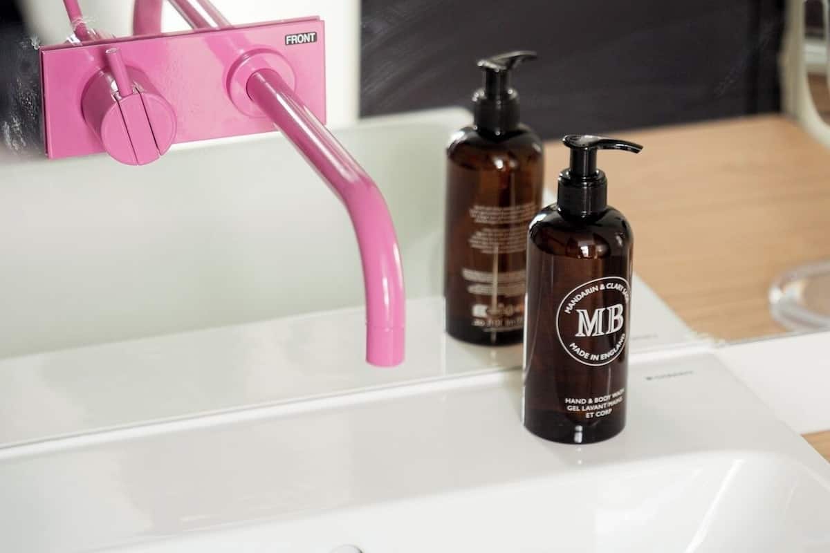 A white bathroom sink with a pink faucet, two brown bottles of hand and body wash, and a mirror.