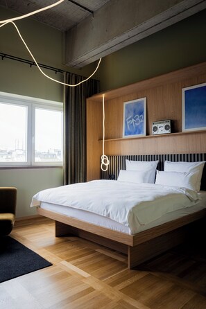 Moderni hotellihuone, jossa on suuri sänky, puinen pääty ja ikkunasta avautuva näkymä kaupunkiin.