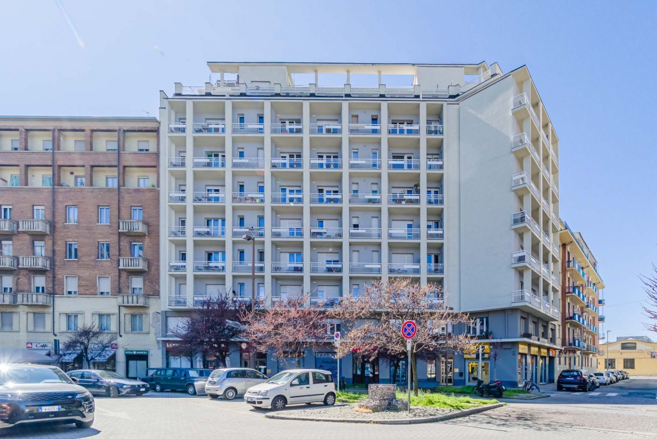 A multi-story residential building with balconies, a parking area, and a small garden with flowering trees.