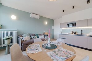 Un salon moderne avec une table à manger pour quatre personnes, une cuisine avec des meubles blancs et un mur gris agrémenté d’un climatiseur.