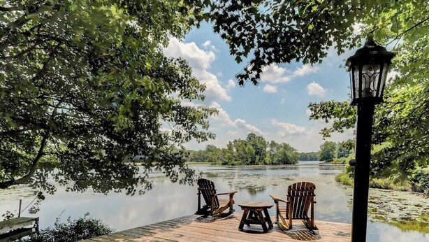 Een houten steiger met Adirondack stoelen en een tafel, met uitzicht op een rustige rivier met bomen aan beide oevers.
