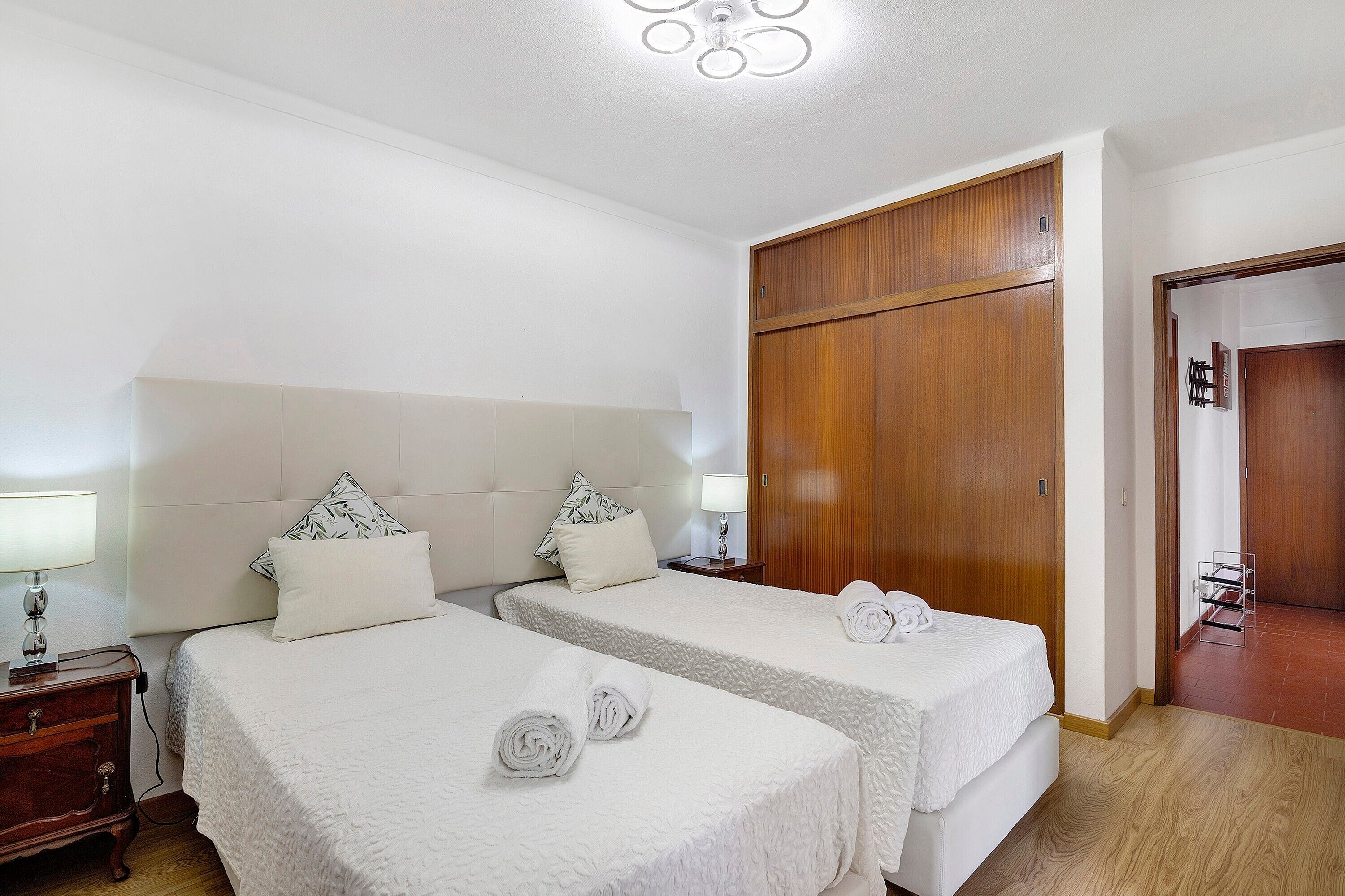 Une chambre d’hôtel avec deux lits, une armoire en bois, une table de chevet avec une lampe et une salle de bain visible à travers une porte ouverte.