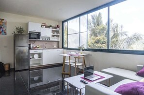 Eine moderne Küche mit Kühlschrank, Mikrowelle und weißen Schränken. Es gibt eine Essecke mit Tisch und Stühlen sowie ein großes Fenster mit Blick auf Palmen.
