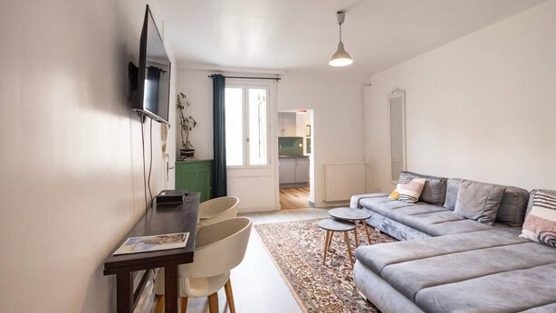 A living room with a sofa, a dining table with chairs, a TV mounted on the wall, and a kitchen area in the background. - The Felix Piyat (Bourges)