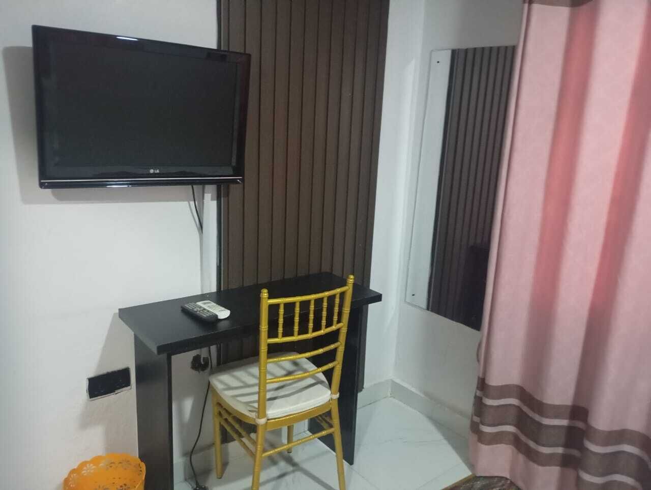 Una habitación pequeña con una televisión montada en la pared, un escritorio con una silla y una cortina entreabierta.
