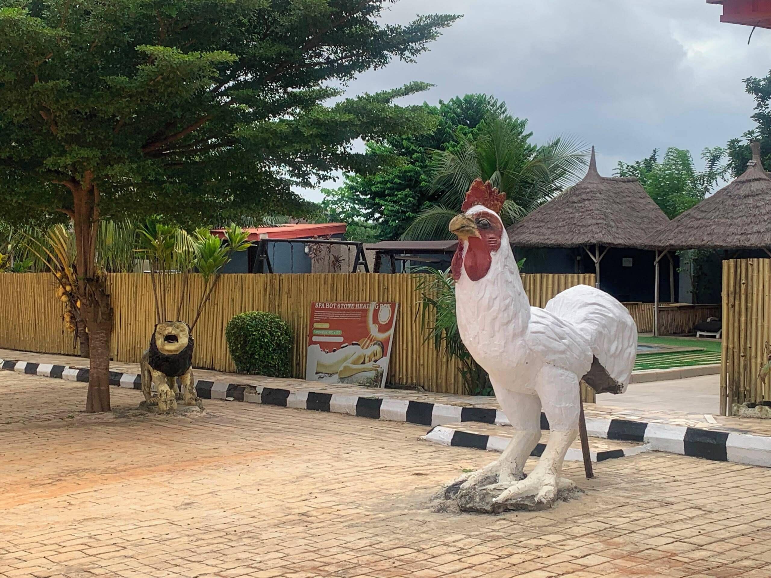 A rooster statue in a courtyard with a bamboo fence, a thatched-roof structure, and a signboard.