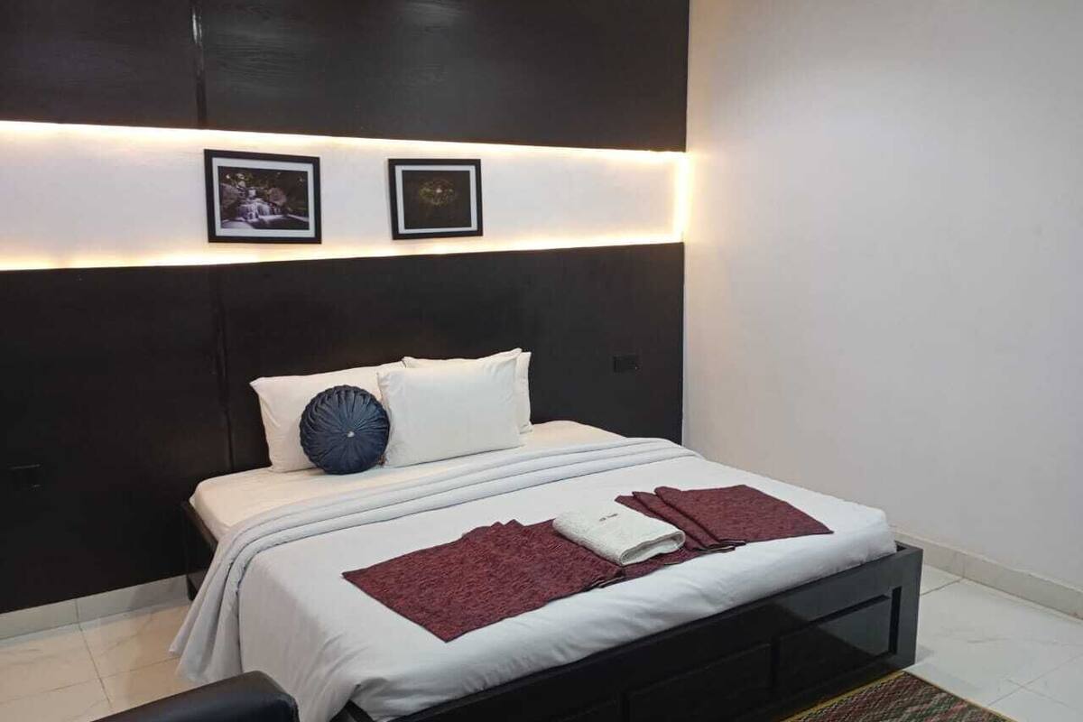 Ein modernes Schlafzimmer mit dunklem Kopfteil, einem Bett mit weißen Bettwäsche, zwei gerahmten Bildern an der Wand und einem Nachttisch mit einer Lampe.