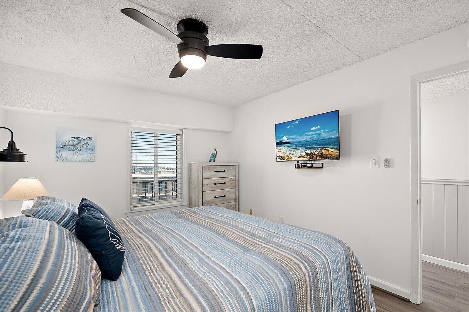 A bedroom with a bed, a ceiling fan, a window with blinds, a dresser, a television, and a wall-mounted picture.