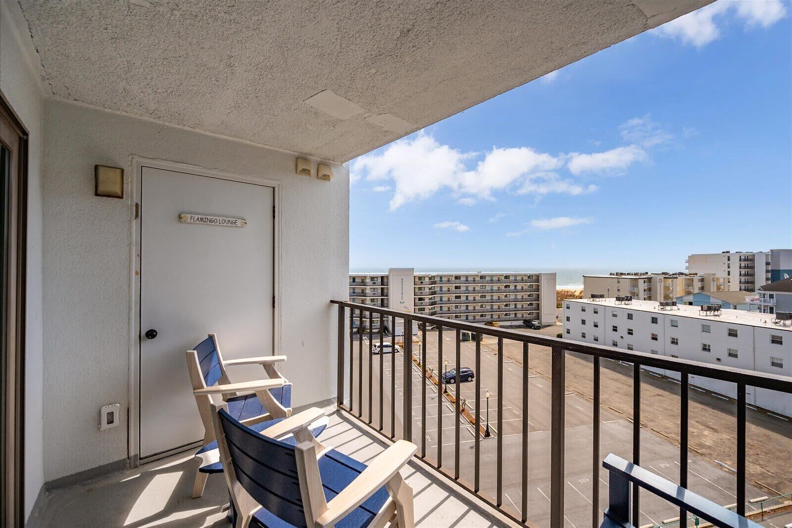 A balcony with a door labeled 'FLAMINGO LOUNGE,' a railing, and a view of a parking lot and buildings.