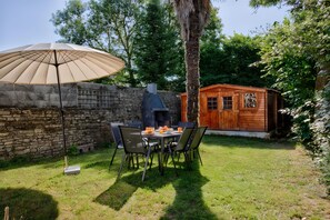 Un mur de pierres, un abri en bois et une table avec des chaises dans une cour arrière verdoyante.