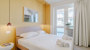 Een hotelkamer met een bed, nachtkastje, lamp, raam en uitzicht op gebouwen.