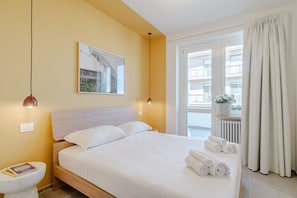 Ein Hotelzimmer mit Bett, Nachttisch, Lampe, Fenster und Blick auf Gebäude.