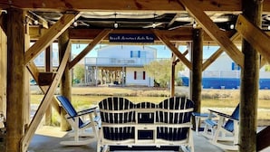 Ein hölzerner Pavillon mit einer Bank und zwei Stühlen, ein Schild mit der Aufschrift 'Beach Memories Here' sowie im Hintergrund ein Haus und ein Strand.