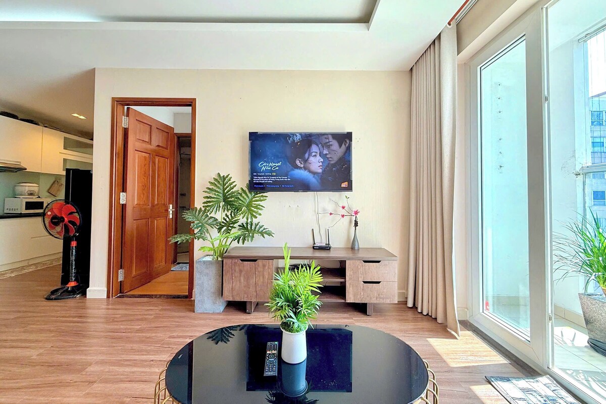 A modern living room with a TV, a wooden door, a plant, a small table, and a large window.
