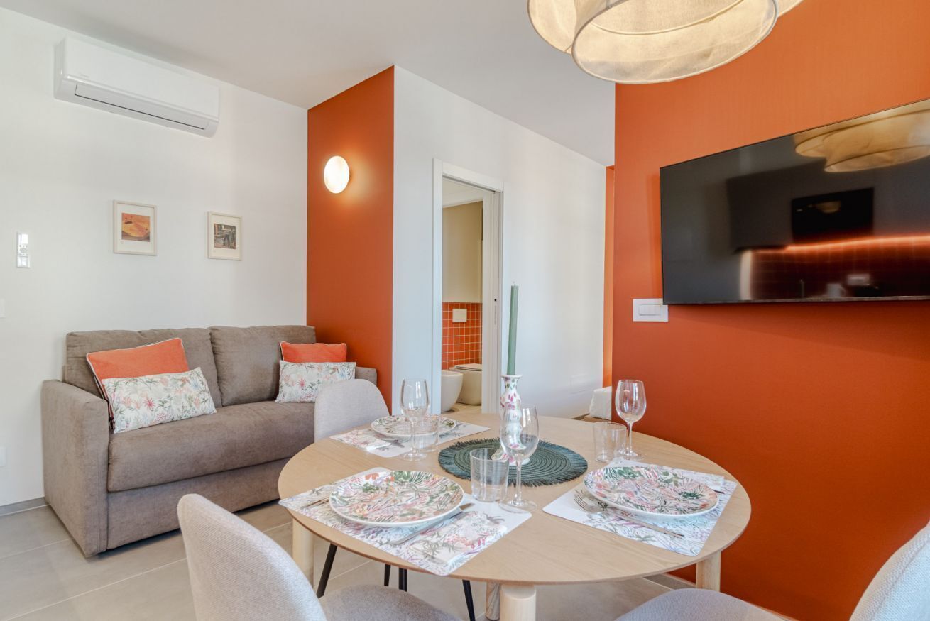A dining area with a round table set for four, a sofa with floral cushions, and a TV mounted on the orange wall.
