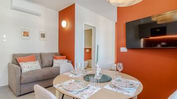A dining area with a round table set for four, a sofa with floral cushions, and a TV mounted on the orange wall.