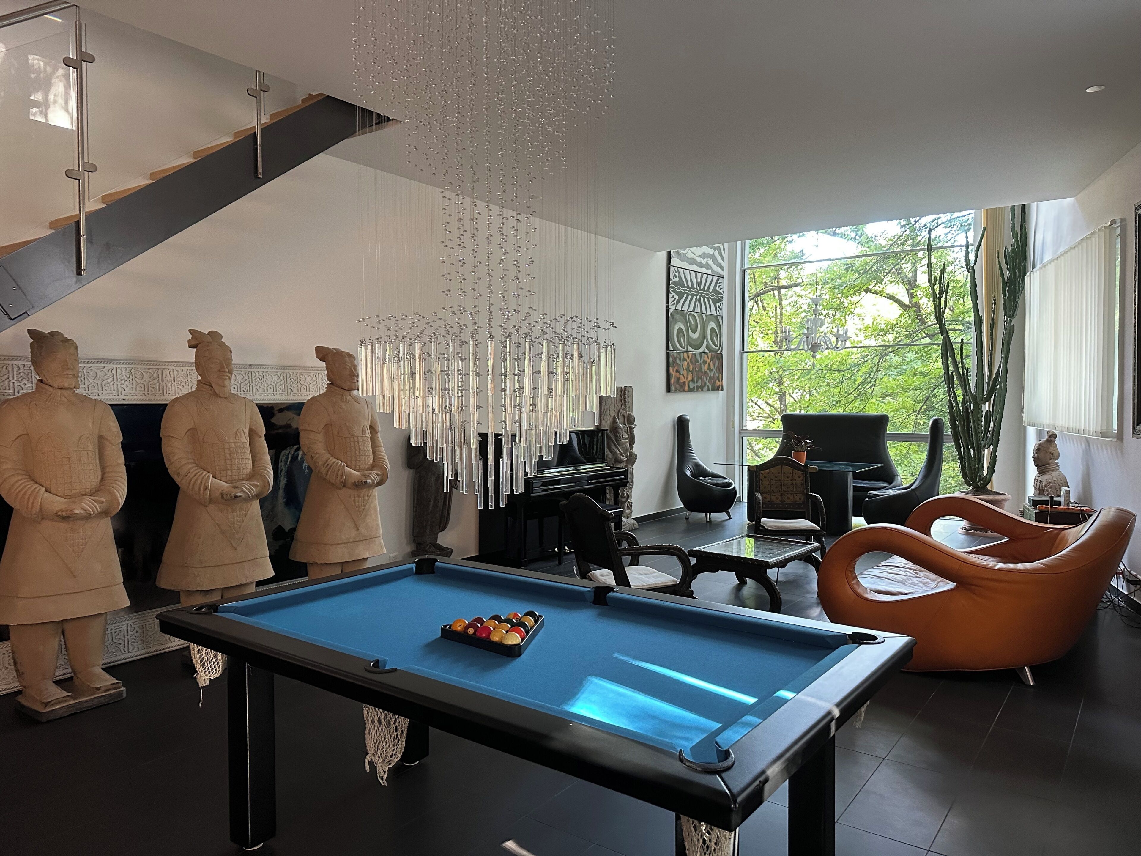 A modern interior with a pool table, two stone statues, a piano, and a large window with a view of greenery.