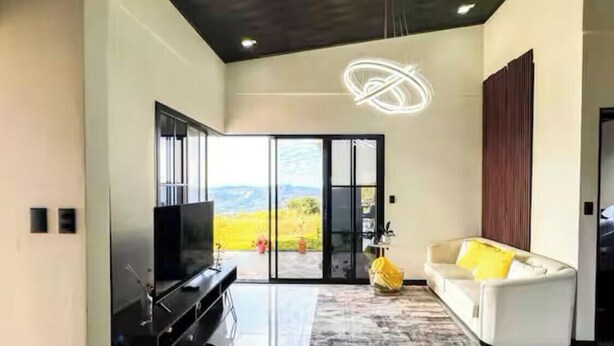 A modern living room with a flat-screen TV, a sofa with yellow cushions, and a large window offering a view of the outdoors. - House Airy Luxury+WiFi+Amazing View At Alajuela CR (Provincia de Alajuela)