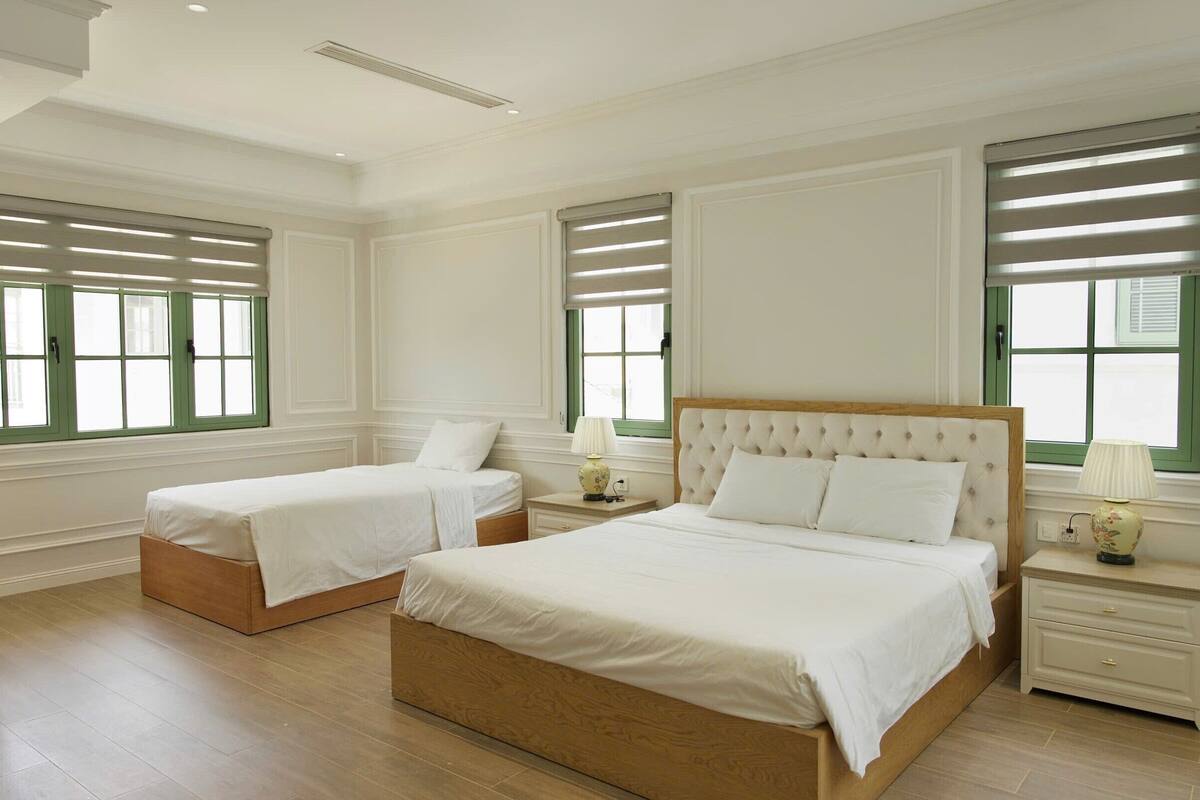 Een hotelkamer met twee bedden, een groot raam met zonwering, een nachtkastje met een lamp en een houten vloer.