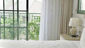 Een slaapkamer met een bed, een nachtkastje met een lamp, een raam met gordijnen en uitzicht op groen buiten.
