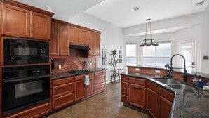 A modern kitchen with wooden cabinets, a black microwave, stove, and oven, a large island with a sink, and a pendant light. - Spacious 4BR Retreat |Pool Table| Firepit & Hottub (Kyle)