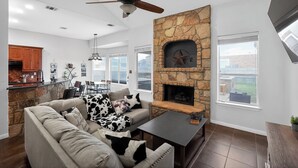A modern living room with a stone fireplace, a ceiling fan, and a dining area with a kitchen in the background. - Spacious 4BR Retreat |Pool Table| Firepit & Hottub (Kyle)