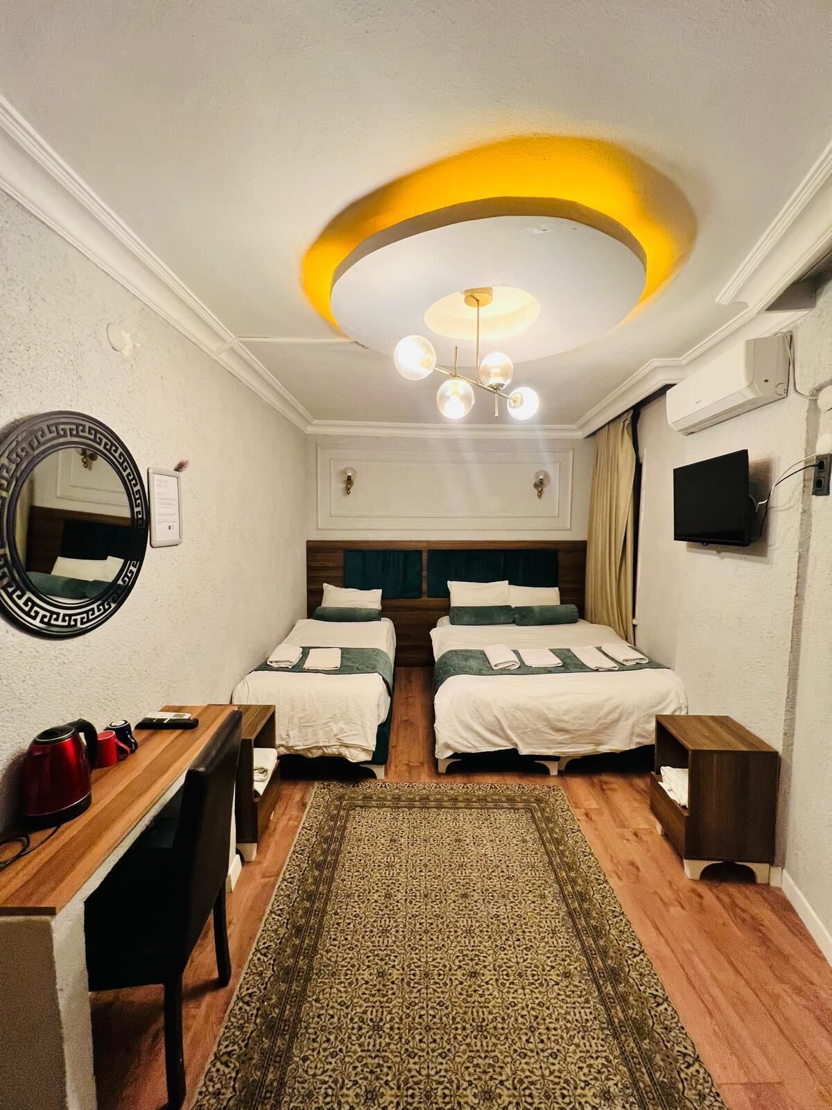 A hotel room with two beds, a desk, a mirror, a TV, and a ceiling light fixture.