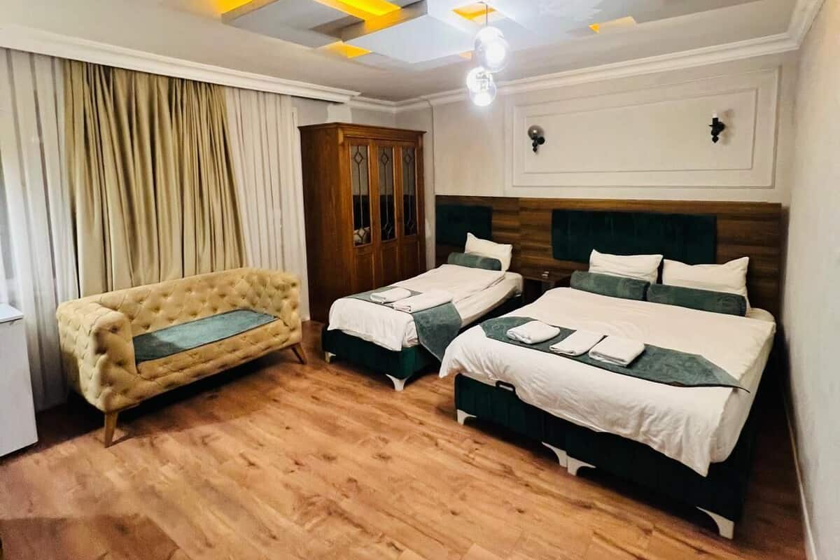 Une chambre d’hôtel avec un plancher en bois, deux lits, un canapé et un plafond orné de luminaires aux formes géométriques.
