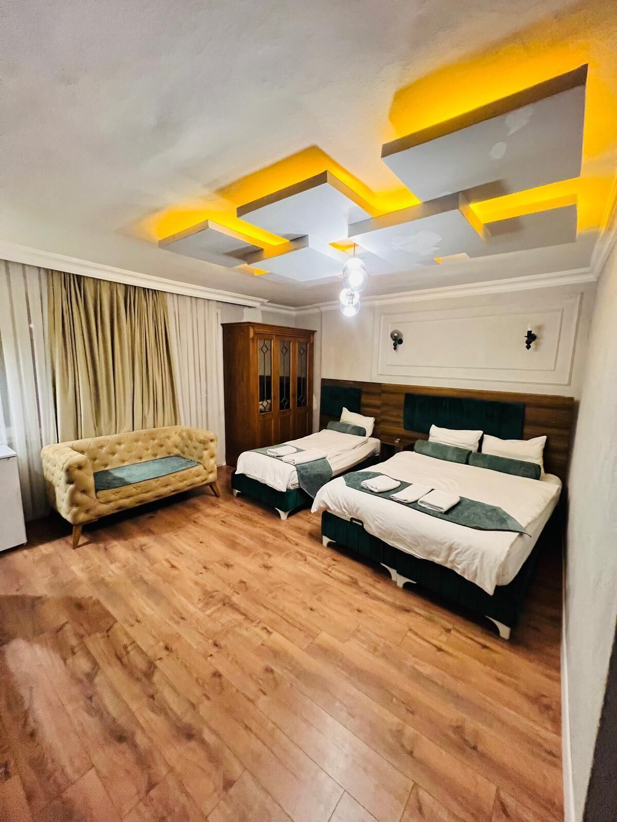 A hotel room with two beds, a sofa, a wardrobe, and a ceiling with geometric light fixtures.