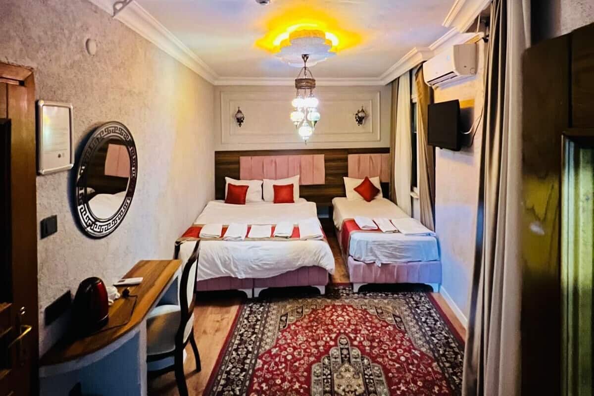 Une chambre d’hôtel avec deux lits, un lustre, un miroir, un bureau et une moquette rouge.