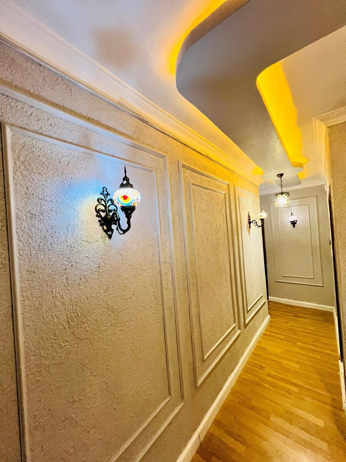 A hallway with decorative wall sconces, wooden flooring, and a curved ceiling with recessed lighting.