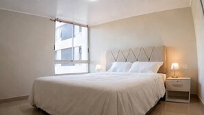Un dormitorio con una cama grande, dos mesitas de noche con lámparas, una ventana y vistas a edificios.
