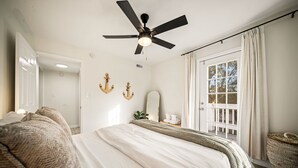 A bedroom with a bed, a ceiling fan, a window with curtains, and a door leading to another room. - NEW! Boho Bungalow (Carolina Beach)